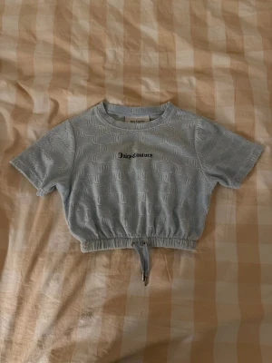 Ljusblå croppad topp Juicy Couture - Säljer en ljusblå croppad topp från Juicy Couture i storlek XXS. Toppen har korta ärmar, broderad logga framtill och ett upphöjt JC-mönster över hela tyget. Den har snörning i midjan för en snygg passform och är gjord i mjuk frotté.