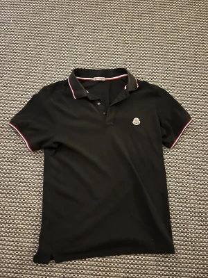 Svart Moncler pikétröja med röd/vit kant - Svart pikétröja från Moncler med klassisk krage och knäppning framtill. Tröjan har röda och vita detaljer på krage och ärmslut samt Moncler-logga på bröstet. Tillverkad i mjuk bomull och har en normal passform. Scan fungerar på pikén.