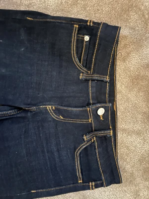 Bootcut jeans från Cheap Monday - Mörkblå bootcut jeans från Cheap Monday med kontrasterande orange sömmar. Klassisk femficksmodell med knapp och dragkedja framtill. Snyggt Cheap Monday-märke på bakfickan. Perfekt för dig som gillar retrovibbar och markerad siluett.