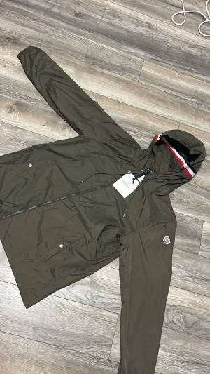 Olivgrön vindjacka från Moncler - Snygg olivgrön vindjacka från Moncler med huva som har röd, vit och marinblå kant. Jackan har dragkedja framtill, snörning vid huvan och två fickor med tryckknappar. Materialet är lätt och syntetiskt, perfekt för blåsiga dagar. HELT NY!