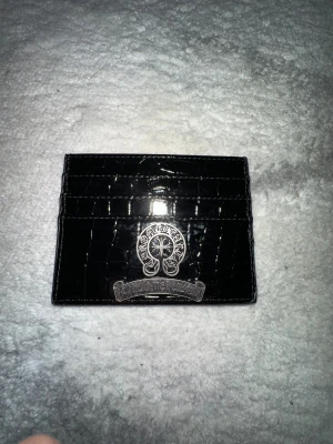 Svart plånbok Chrome Hearts - Snygg svart plånbok från Chrome Hearts med croco-mönstrad yta i skinn och en stor silvrig metallapplikation framtill. Plånboken har flera fack för kort och är perfekt för dig som gillar exklusiva detaljer och stilren design.