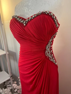 Klänning - Röd långklänning med axelbandslös design och hjärtformad urringning. Klänningen har draperad framsida och är dekorerad med glittrande strass längs urringningen och sidan. Använd en gång och i utmärkt skick ❤️