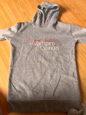 Grå Vampire Diaries hoodie XS - Grå hoodie med huva och tryck 'The Vampire Diaries' på bröstet. Långärmad med två vita ränder på båda ärmarna. Mjuk insida och ribbad nederkant. Perfekt för dig som älskar serien och vill ha en chill look.