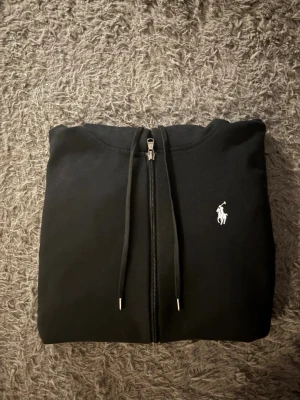 Svart hoodie från Polo Ralph Lauren - Svart hoodie med dragkedja från Polo Ralph Lauren. Klassisk design med huva och snörning, vit broderad logga på bröstet och två fickor framtill. Tillverkad i mjuk bomull, perfekt för chill dagar. Långärmad och loose fit för skön komfort. Rekommenderar att man är ca 170 lång