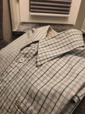 Rutig skjorta från Blend of America, storlek L - Klassisk långärmad skjorta från Blend i storlek L. Skjortan är vit med gröna och grå rutor, har knappar framtill och en bröstficka med broderad logga. Tillverkad i bomull och har en snygg krage med kontrastmönster på insidan.