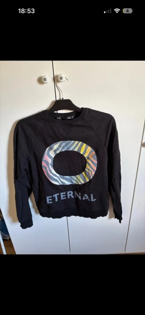 Svart sweatshirt från 157 med tryck - Cool svart sweatshirt från 157 i storlek M med stort färgglatt O-tryck på bröstet och texten 'ETERNAL' under. Trycket har zebra-inspirerade ränder i blått, gult, rosa och grått. Tröjan har rund hals och långa ärmar, perfekt för dig som gillar streetstyle.