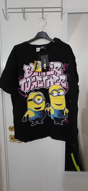 Svart Minions t-shirt från FB Sister - Cool svart t-shirt från FB Sister med Minions-tryck och texten 'Better Together' i rosa graffiti-stil. Loose fit och kortärmad modell, perfekt för dig som gillar roliga prints och streetwear-vibe. Materialet är mjuk bomull för skön känsla hela dagen.