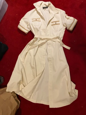 beige 50-talsklänning - retro beige skjortklänning. med knappar hela vägen, kort ärm och bälte i midjan. två bröstfickor med knappdetaljer. 50-tal förmodligen, köpt i tyskland. passar S-M