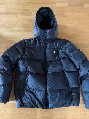 Svart pufferjacka från Calvin Klein - Snygg svart pufferjacka från Calvin Klein med huva och två praktiska fickor med dragkedja. Jackan har CK-logga på bröstet och är perfekt för kalla dagar. Materialet är matt/glansig och känns riktigt modernt. Passformen är normal. Använd fåtal gånger priset är ej hugget i sten