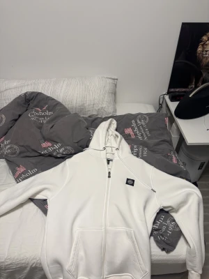 Vit hoodie från Stone Island, storlek L - Vit hoodie från Stone Island med klassisk logopatch på bröstet och ärmen. Tröjan har huva med snörning, dragkedja framtill och två stora fickor. Tillverkad i mjuk bomull som är skön mot huden. Perfekt för dig som gillar clean och stilren streetwear. Den håller dig varm också om de kallt ute. Aldrig användas. 