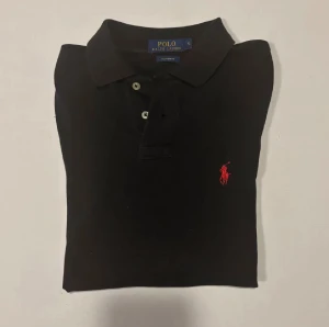Svart pikétröja från Ralph Lauren - Svart pikétröja från Ralph Lauren med klassisk krage och två knappar framtill. Tröjan har det ikoniska röda Polo-logot broderat på bröstet. Tillverkad i mjuk bomull som är skön mot huden och har en snygg, normal passform.