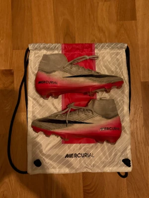 Nike Mercurial Air Zoom fotbollsskor - Säljer ett par Nike Mercurial Air Zoom fotbollsskor i storlek 41. Skorna har snörning, hög strumpliknande krage och platt sula med dobbar för gräsplan. Skorna är använda ett fåtal gånger och är i bra skick. Materialet är syntet och skinn, med en snygg gradient från grått till rött och Nike-logga på sidan. Pris kan diskuteras