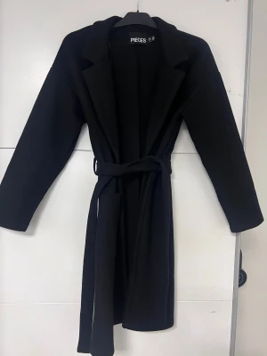 Svart kappa - Svart kappa med klassisk krage, långa ärmar och två stora fickor framtill. Modellen är rak med bälte. 