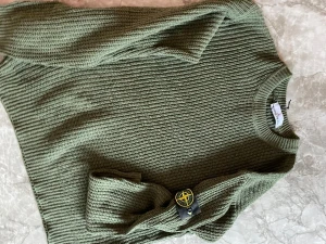 Grön stickad Stone Island tröja - Snygg grön stickad tröja från Stone Island med klassisk rund hals och ribbade muddar. Tröjan har lång ärm och den ikoniska Stone Island patchen på ena ärmen. Perfekt för dig som gillar streetwear och vill ha en chill look.