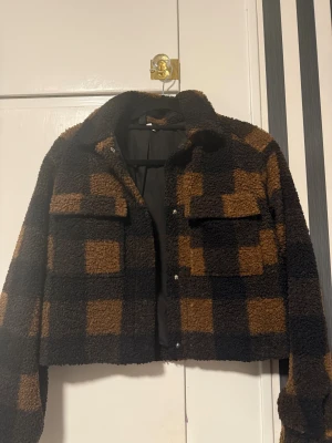 Rutig teddyjacka från H&M  - Säljer en croppad teddyjacka i storlek XS. Jackan är brun och svart med rutigt mönster, har två bröstfickor och stängs med knappar framtill. Perfekt för dig som vill ha en mysig och trendig look.