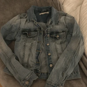 Ljusblå jeansjacka med knappar - Snygg ljusblå jeansjacka från Bik Bok med klassisk krage, långa ärmar och silverfärgade knappar framtill. Jackan har två bröstfickor med lock och knapp, samt dekorativa sömmar som ger en cool vintagekänsla. Perfekt att slänga över en hoodie eller t-shirt.