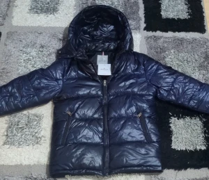 Mörkblå dunjacka från Moncler - Snygg mörkblå dunjacka från Moncler med glansig finish och huva. Jackan har två sidofickor med dragkedja, quiltad design och klassisk Moncler-logga på insidan. Perfekt för kalla dagar och har en modern look som passar streetstyle.