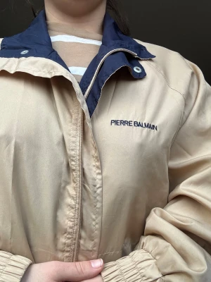 Pierre Balmain beige vindjacka - Pierre Balmain vindjacka i beige med marinblå detaljer och krage. Jackan har dragkedja framtill, elastiska muddar och logga broderad på bröstet. Snygg loose passform och tillverkad i lätt polyester som funkar perfekt när det blåser.