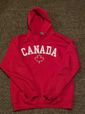 Röd hoodie med Canada-tryck - Säljer en röd hoodie från Gildan med stor vit text 'CANADA' och ett lönnlöv på bröstet. Tröjan har huva, magficka och långa ärmar. Perfekt för dig som gillar streetwear och vill sticka ut med en bold look.