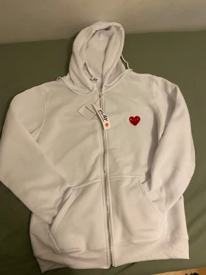 Vit hoodie från Comme des Garçons Play - Vit hoodie från Comme des Garçons Play med dragkedja och huva. På bröstet finns det ikoniska röda hjärtat med ögon broderat. Tröjan har långa ärmar, fickor framtill och ribbade muddar. Perfekt för dig som gillar stilrena och trendiga plagg.