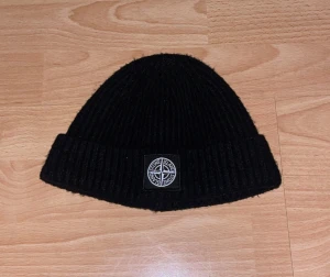 Svart Stone Island mössa  - Svart ribbstickad mössa från Stone Island med klassisk logopatch framtill. Tillverkad i mjuk ull och har en uppvikt kant för extra stilpoäng. Perfekt för dig som vill ha en clean och snygg look under kalla dagar.