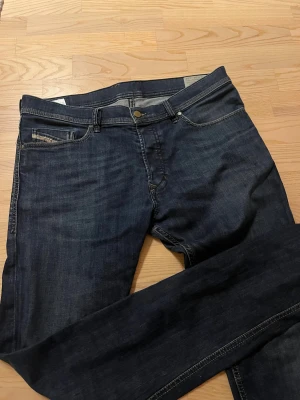 Diesel mörkblå jeans herr - Snygga Diesel jeans i mörkblå tvätt med klassisk femficksdesign och smal passform. Jeansen har diskreta slitningar, silverfärgade detaljer och logopatch bak.