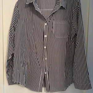 Svartvit randig skjorta med knappar - Snygg skjorta med svartvita vertikala ränder och klassisk krage. Skjortan har långa ärmar, en bröstficka och vita knappar framtill. Perfekt för dig som gillar stilrena och grafiska mönster. Materialet känns lätt och skönt mot huden.