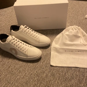 Oliver Cabell  - Snygga vita sneakers från Oliver Cabell med rund tå och platt sula. Skorna är tillverkade i slätt skinn med svart hälparti och en beige cirkeldetalj på sidan. Klassisk snörning och stilren design, perfekt för dig som gillar minimalistiska och trendiga sneakers. Helt nya aldrig använda. Både box och påse ingår i köpet. Skriv vid minsta fundering.