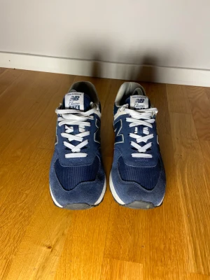 New Balance 574 Classic blå sneakers - Säljer ett par New Balance 574 Classic sneakers i marinblått med vita detaljer och snörning. Skorna har en platt sula och är tillverkade i mocka och syntetmaterial. Strl 44 | Skick 8,5/10 | (Orginal snören medföljer) 