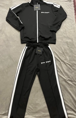 Palm angels dress - Palm Angels svart zip-dress med vita ränder längs ärmarna och logga på bröstet. Tröjan har ribbade muddar, ståkrag och hel dragkedja framtill. Materialet är mjukt och sportigt, perfekt för en chill och trendig look.