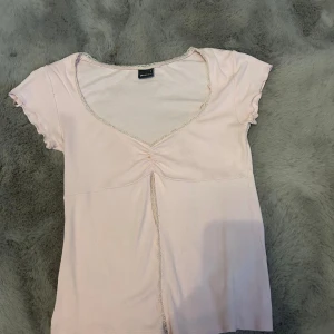 Ljusrosa v-ringad topp med spets - Söt ljusrosa topp från Gina tricot med v-ringning och spetsdetaljer längs halsringningen och framtill. Toppen har korta ärmar med vågig kant och en liten rynkdetalj vid bysten. Perfekt för en trendig och feminin look.