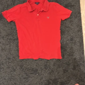 Röd pikétröja från Gant XL - Snygg röd pikétröja från Gant i klassisk modell med krage och två knappar framtill. Tröjan har broderad Gant-logga på bröstet och är tillverkad i mjuk bomull. Perfekt för en stilren och avslappnad look.