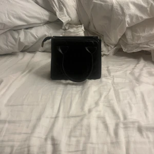 Svart liten handväska i - Snygg svart handväska med stilren, fyrkantig form och praktiskt handtag. Väskan har dragkedja upptill med gulddetaljer och ljus insida. Perfekt storlek för det viktigaste och ger en cool vibe till din outfit.