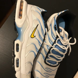 Nike Air Max Plus vita/blå sneakers Tn - Nike Air Max Plus TN sneakers i vitt med blå detaljer och svarta inslag. Swoosh-loggan är gul och svart. Skorna har en mesh-överdel, synlig Air Max-dämpning i sulan och snörning framtill. Designen har vågiga linjer och en sportig vibe, perfekt för dig som gillar streetwear. Mycket bra skick använda max 2-3 gånger