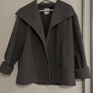 Mörkgrå kappa med bred krage - Snygg mörkgrå kappa med bred krage och stora knappar framtill. Jackan har uppvikta ärmslut och en loose passform som ger en chill vibe. Perfekt för dig som gillar stilrena och enkla plagg med lite edge. Materialet känns som ull och är mjukt och varmt.