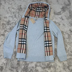 Grå Burberry hoodie med klassiskt rutmönster - Snygg grå hoodie från Burberry med dragkedja och huva. Insidan av huvan har det ikoniska beigea rutmönstret. Broderad Burberry-logga på bröstet och praktiska fickor framtill. Perfekt för dig som vill ha en stilren och lyxig streetwear-look.