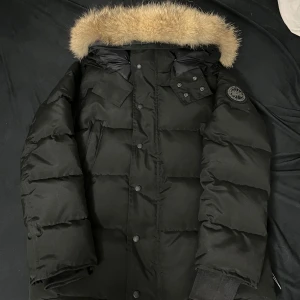 Svart dunjacka från Canada Goose - Svart dunjacka från Canada Goose med fluffig beige päls på huvan, patch på ärmen och flera fickor framtill. Jackan har en quiltad design, dragkedja och knappar. Perfekt för kalla vinterdagar med sin tjocka och varma fyllning. VID MER FRÅGOR ELLER BILDER ÄR DET BARA ATT SKICKA PRISET KAN SPNKAS VID SNABBA AFFÄRER 