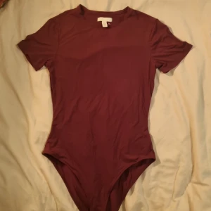 Vinröd bodysuit från H&M, strl S - Säljer en vinröd bodysuit från H&M i storlek S. Modellen har korta ärmar, rund hals och knäppning i grenen. Materialet är mjukt och stretchigt, perfekt för att styla med jeans eller kjol. Enkel och clean design utan mönster eller detaljer.