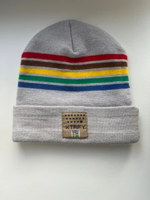 Randig grå mössa från Xtrfy - Snygg grå mössa med bred uppvikt kant och broderad Xtrfy-patch framtill. Mössan har coola horisontella ränder i rött, brunt, gult, grönt och blått. Materialet är stickad syntet. Texten 'PRESS START' är broderad i vitt på kanten. Limiterad upplaga som ej går att köpa. Finns fläckar (se bilder) som går att tvätta bort.