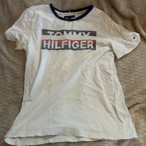 Vit Tommy Hilfiger t-shirt med tryck - Vit t-shirt från Tommy Hilfiger med klassiskt logotryck i blått och rött på bröstet. Rund hals med mörkblå kant och korta ärmar. Perfekt för en chill och stilren look, ikonisk design med broderad logga på ärmen.