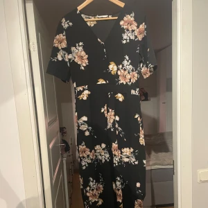 Svart blommig byxdress från ONLY - Svart byxdress från ONLY med v-ringning och korta ärmar.  Blommigt mönster i beige, rosa och vitt, samt markerad midja med lätt rynkning. Tillverkad i mjuk polyester som faller snyggt och har knappar framtill. Obs, mer som en Medium än Large