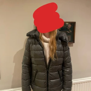 Mörkgrå Moncler pufferjacka med huva - Säljer en snygg mörkgrå pufferjacka från Moncler med broderad logga på ärmen. Jackan har huva, dragkedja framtill och två fickor med dragkedja. Materialet är glansigt och känns riktigt lyxigt, perfekt för kalla dagar. Passformen är normal och jackan är långärmad. Den har lite små defekter, därav priset. Kan skicka bilder om du vill det. Kontakta så skickar jag äktenhetsbevis. Tryck ej på ”köp nu” om jag inte svarat dig än. 