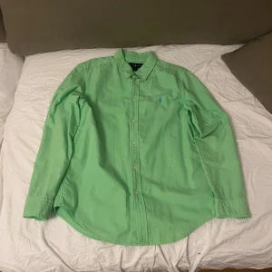 Grön skjorta från Ralph Lauren  - Fräsch grön skjorta från Ralph Lauren i storlek XL 18-20 vilket motsvarar medium i vanlig storlek. Klassisk button-down krage, långärmad och med knappar hela vägen framtill. Skjortan har den ikoniska blå Ralph Lauren-loggan broderad på bröstet. Tillverkad i mjuk bomull som känns skön mot huden.