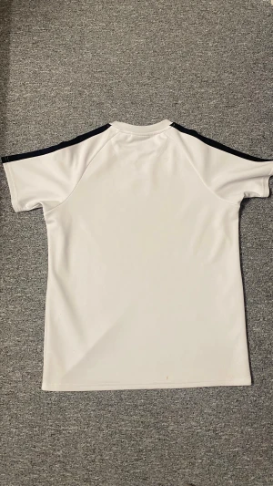 Vit McKenzie t-shirt med svarta detaljer - Snygg vit t-shirt från McKenzie med svarta paneler längs axlarna och röda sömmar som ger en sportig vibe. T-shirten har rund hals, korta ärmar och är gjord i mjuk polyester som andas. Perfekt för dig som gillar stilren och modern sportwear.