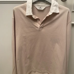 Pikétröja - Använd 2 gånger. Priset kan diskuteras. Snygg beige pikétröja från H&M med vit klassisk krage och två knappar vid halsen. Tröjan är långärmad och har en relaxed passform. Tillverkad i 100% bomull för en skön och mjuk känsla. Perfekt för dig som gillar stilrena och enkla plagg.