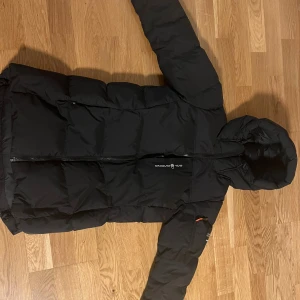 Svart Sail Racing pufferjacka dam  - Svart pufferjacka från Sail Racing med huva och snyggt Sail Antarctica Expedition-märke på ärmen. Jackan har lång dragkedja framtill, quiltad design och är tillverkad i syntetmaterial. Perfekt för kalla vinterdagar och har en clean, sportig look.