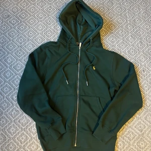 Mörkgrön hoodie från Polo Ralph Lauren - Snygg mörkgrön hoodie från Polo Ralph Lauren med gul  logga . Tröjan har huva med snörning, dragkedja framtill och en stor magficka. Tröjan är i nyskick och det förklarar lappen kvar. Storlek medium. Hör svenska f vid några frågor.