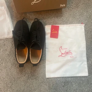 Svarta sneakers från Christian Louboutin - Säljer ett par svarta sneakers från Christian Louboutin med klassisk röd sula och snörning. Skorna har en rund tå och är tillverkade i mocka med skinnfoder. Kommer med originalpåse och box. Perfekt för dig som gillar exklusiva och stilrena sneakers. Box,skoskydd, kort ingår! Skriv vid fundering. Finns en liten skada på ovansidan som inte syns vid användning