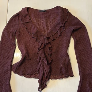 Vinröd volangblus från Gina Tricot - Supersöt vinröd blus från Gina Tricot med volangdetaljer längs v-ringningen och framtill. Långärmad modell med utsvängda ärmslut och lätt transparent struktur. Perfekt för dig som gillar romantiska vibes och snygga detaljer.