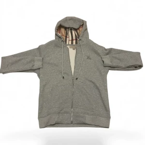 Grå hoodie från Burberry med dragkedja - Säljer en grå hoodie från Burberry med klassiskt rutigt foder i huvan och broderad logga på bröstet. Tröjan har långa ärmar, snörning i huvan och dragkedja framtill. Perfekt för dig som gillar stilren design med lyxig touch.